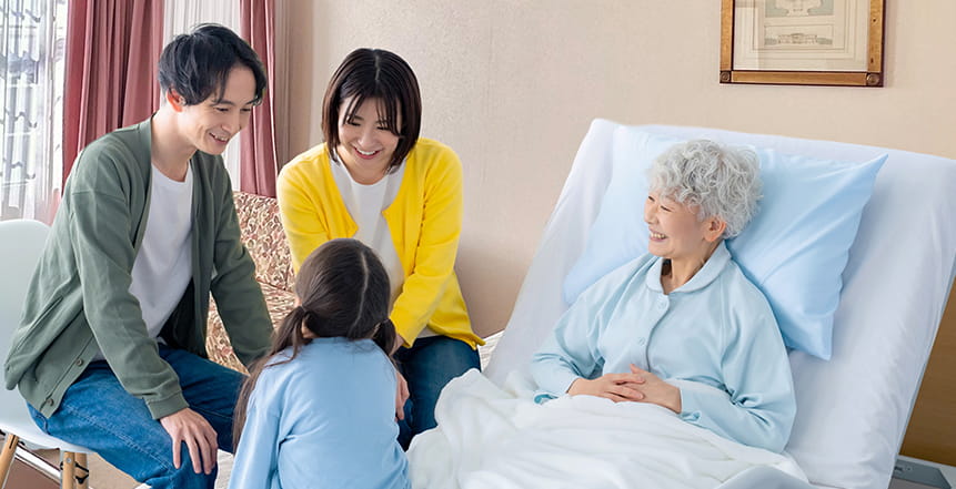 病室でベッドに座っている女性を3人の家族が見舞いに来ている様子